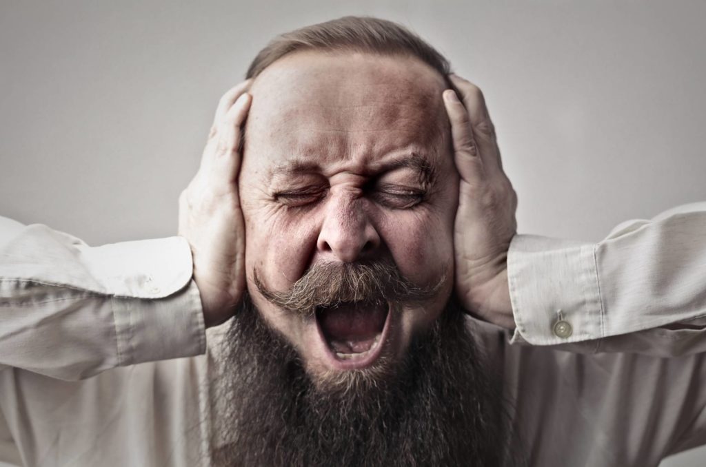 close up photo of screaming man with a full beard covering his ears and closing his eyes. Maybe he needs to goof off?