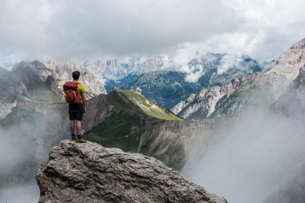 man with red backpack standing on cliff facing mountains under white sky during daytime